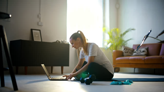 SLOW MOTION Fit young woman sitting and using laptop on yoga mat,preparing for online home workout on sunny living room floor. Healthy lifestyle,exercise,fitness,active lifestyle,domestic life. Shot in 8K Resolution.
