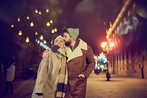 Young happy woman enjoying in a kiss by her boyfriend while walking embraced during Christmas night on the city street.