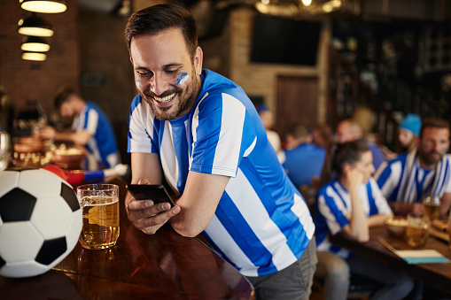 Happy soccer fan text messaging over smart phone before watching a game in a bar.