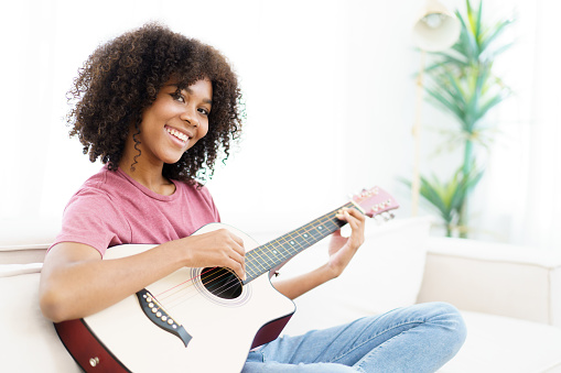 Happy cheerful American - African black woman sitting on a sofa in living room and playing an acoustic guitar close up with copy space.