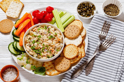 healthy easy leftover salmon salad served with fresh carrots, cucumber, celery, tomatoes and crackers on white plate on white wood table, horizontal view from above