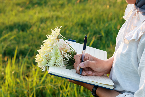 portrait of woman writing in a book with a bunch of beautiful flowers. woman in hijab writing while standing outdoors