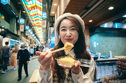 From a second-person perspective, an Asian woman is traveling with friends and family in Nishiki Market in Kyoto, Japan. They are together indulging in and sharing the local cuisine.
