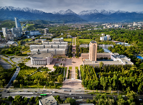Aerial drone view panorama of Kazgu university educational center square near timiryazev street with cars and buildings around in Almaty city against snow mountains at background in Kazakhstan