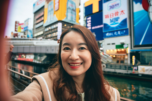 Through a selfie perspective, an Asian woman is capturing her travel memories in Dotonbori, Osaka, and live streaming to share with friends and family on social media.