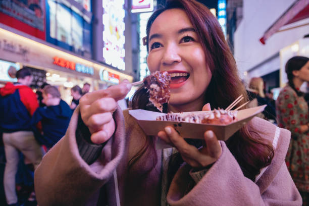 A female tourist in Osaka is holding a freshly made Japanese traditional street food called Takoyaki (octopus balls) as she explores the city center streets. We can see her hand holding the piping hot Takoyaki skewer, wafting with a tantalizing aroma.The bustling city streets serve as the backdrop, filled with vibrant shops, colorful signage, and a multitude of people moving about. The sounds of chatter and street performers create an energetic atmosphere. una turista en osaka sostiene una comida callejera tradicional japonesa recién hecha llamada takoyaki (bolas de pulpo) mientras explora las calles del centro de la ciudad. podemos ver su mano sosteniendo la brocheta takoyaki muy caliente, flotando con un