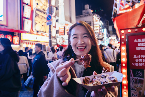 A female tourist in Osaka is holding a freshly made Japanese traditional street food called Takoyaki (octopus balls) as she explores the city center streets. We can see her hand holding the piping hot Takoyaki skewer, wafting with a tantalizing aroma.The bustling city streets serve as the backdrop, filled with vibrant shops, colorful signage, and a multitude of people moving about. The sounds of chatter and street performers create an energetic atmosphere.