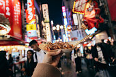 A female tourist in Osaka is holding a freshly made Japanese traditional street food called Takoyaki (octopus balls) as she explores the city center streets. With the camera angled from her personal perspective, we can see her hand holding the piping hot