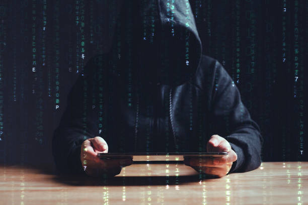 Black hat hacker using tablet on table where sensitive data is hacked in a dark room background with matrix binary rain code. Cyber security and cyber crime concept. Hacking and phishing stock photo