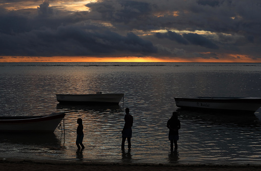 Albion, Mauritius - April 26, 2023: Silhouettes of angler and boats at the beach of the Indian Ocean in the West of Mauritius during sunset.