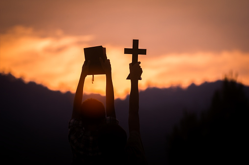 Two children holding christian cross and bible with light sunset background.