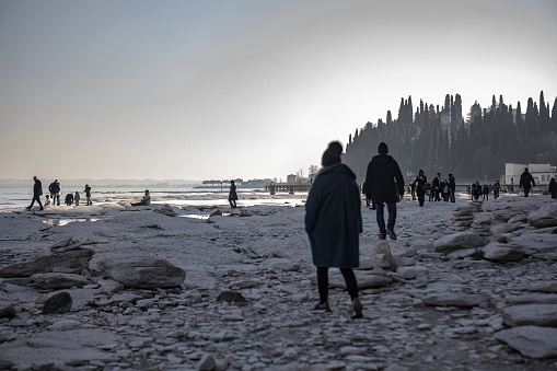 Sirmione, Italy 15 February 2023: A nostalgic shot in black and white, featuring people walking on a rocky shoreline by a lake. The dreamy atmosphere of the photo evokes a sense of longing for simpler times.