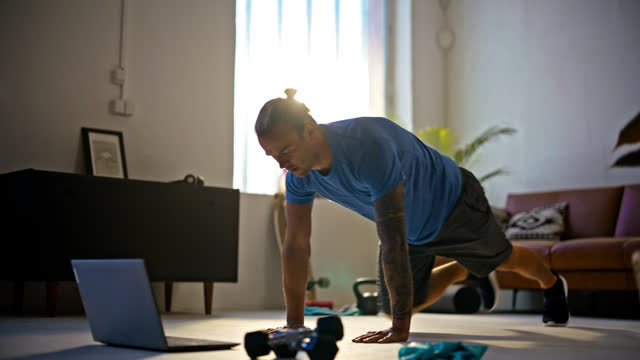 SLO MO Fit young man with tattoos and man bun doing mountain climber exercises at laptop on living room floor,doing home workout. Healthy lifestyle,exercise,fitness,active lifestyle,strength. Exercising early in the morning.