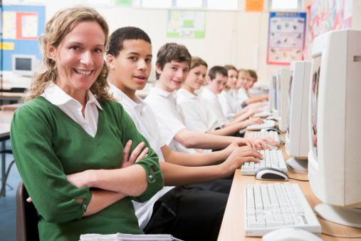 Row of schoolchildren studying in front of a computer with teacher smiling to camera