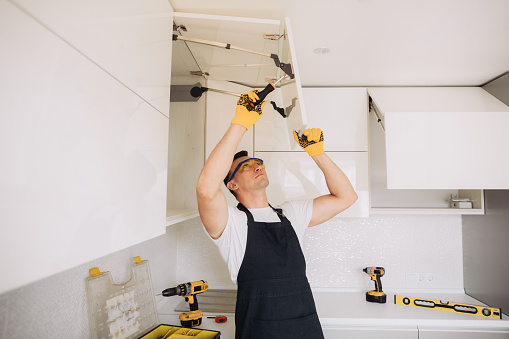 Maintenance man installing kitchen furniture in a black apron and yellow gloves using a power skrewdriver