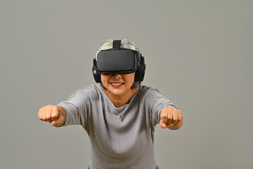 Cheerful senior woman with virtual reality glasses simulating of driving car in VR glasses isolated on white background.