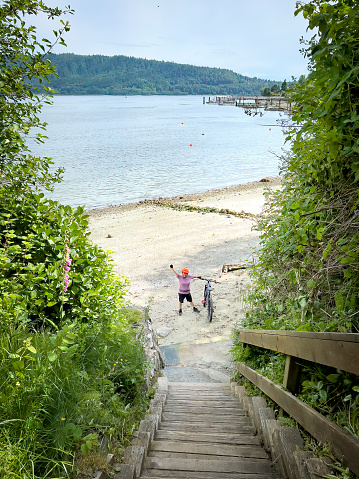A senior Chinese woman mountain biker gives a fist pump after descending a long flight of stairs to discover a sandy deserted beach.  Deep Cove, North Vancouver, British Columbia, Canada