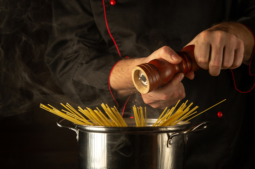 The chef adds pepper to a hot pot of spaghetti. Free space for recipe on dark background.