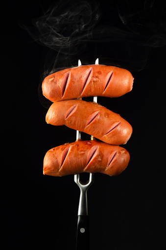 Grilled hot with steam three sausages on a fork. Tasty and fast food concept on black background