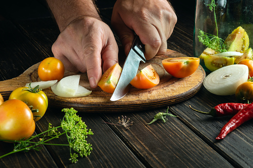 The chef cuts tomatoes with a knife on a kitchen cutting board for canning in a jar. Diet food by the hands of the cook in the kitchen