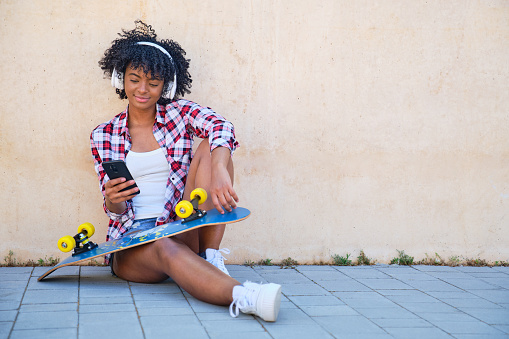young afro girl sitting on the floor with her skateboard on her legs listening to music with her smart phone and white headphones