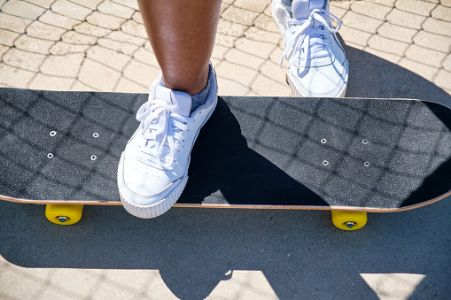 image from above of the feet of an African-American girl stepping on a skateboard with white sneakers with the shadow of a metal fence in the background