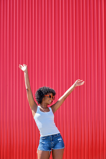 smiling african american girl in orange sunglasses with her arms up on a red background with plenty of copy space above
