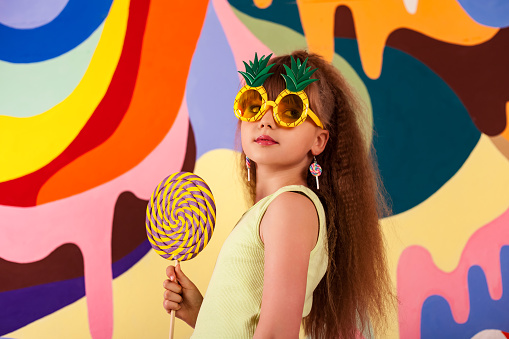Little girl holding round giant lollipop at colored wall, looking side away. Kid 9-10 year old in yellow t-shirt and pineapple sunglasses with candy on stick. Summer fashion concept. Copy text space