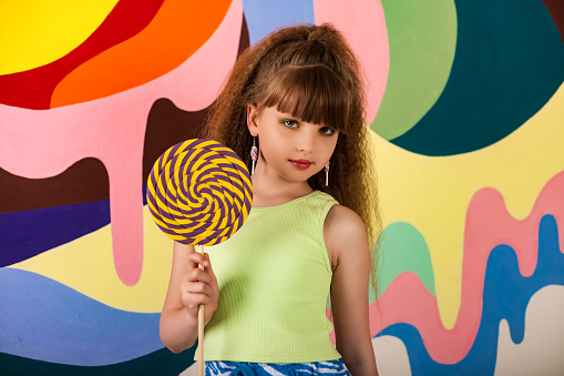 Charming little girl holding round giant lollypop at colored wall, looking at camera. Kid 9-10 year old in yellow t-shirt and hairstyle with candy on stick. Summer fashion concept. Copy ad text space