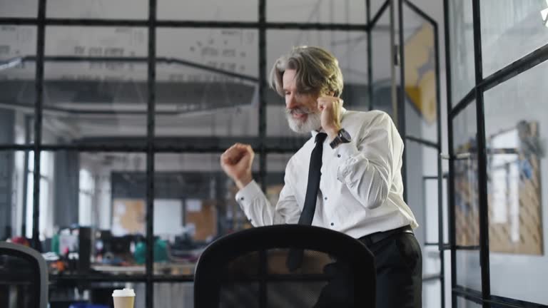 Happy nature businessman with grey hair and beard in formal clothes is in the office celebrating success and dancing