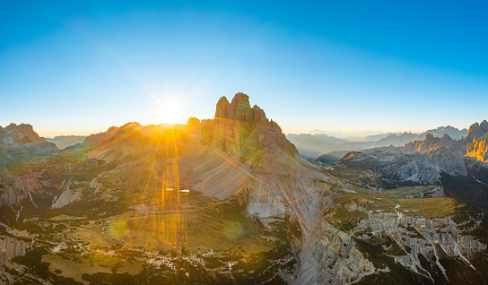 Sun rises over mountain range. Rocky mountains with sand-covered foothills and green valley. Three Peaks of Lavaredo at sunrise aerial view in back lit