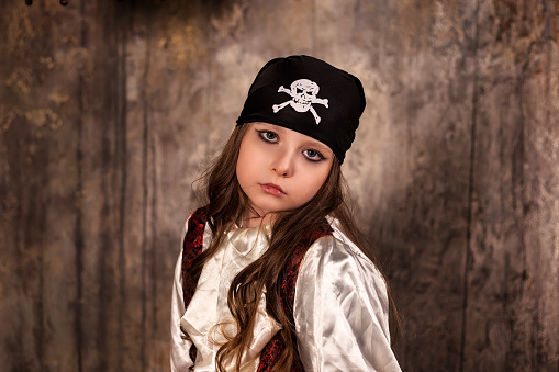 Portrait of pensive little girl actress 7-8 year old in pirate's image at grey textured wall, looking at camera. Actor child posing pirate corsair costume on stage. Theatre style concept. Copy space