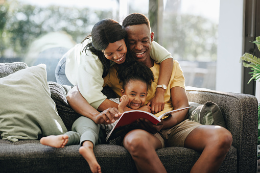 Parents reading a book with their daughter on a couch. Happy family spending time together at home. African couple raising a toddler in a loving environment.