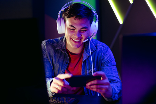 Happy video game player watching a live stream of a gameplay on his smartphone. Male gamer sitting at his gaming station with headphones on, enjoying an exciting gaming broadcast on a mobile streaming app.