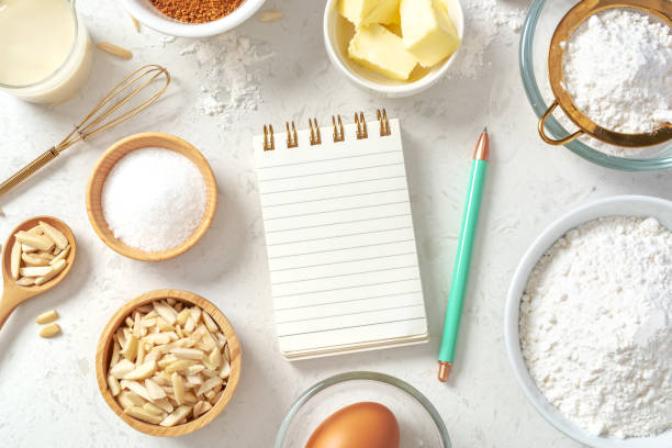 Notepad and pen for recipe with baking or cake ingredients in bowls, butter, milk, nuts and eggs with gold whisk on marble table