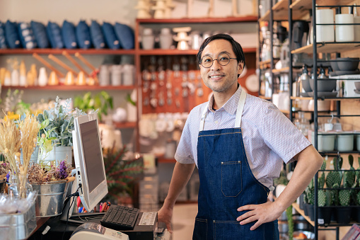 Good service makes business grow steadily. Smart Japanese owner standing at cashier counter receive order from customer looking at camera and smile at the home decoration shop in the city. Shopping and service concept.