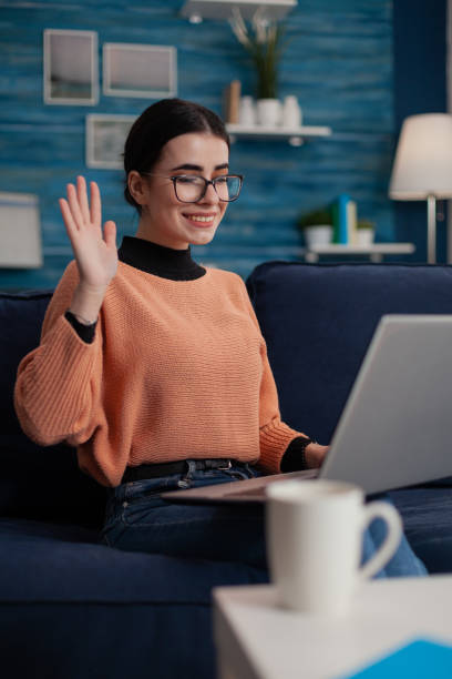 istock Smiling student on sofa with laptop on lap waving hello in videocall. 1493473519