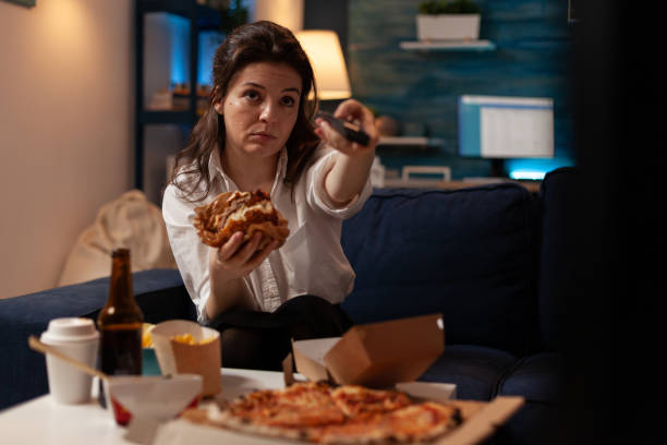istock Young woman sitting on sofa eating double cheeseburger holding remote turning up volume 1493474886
