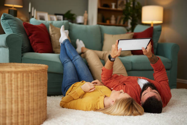young couple using a tablet while relaxing on carpet at home - couple loving lying on back carpet imagens e fotografias de stock