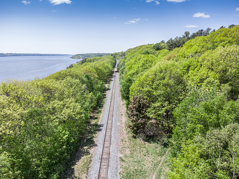 Aerial view of railroad surrounded by trees and besides the St. Lawrence river during a day of summer in Quebec city.\nThe train is coming from the distance.
