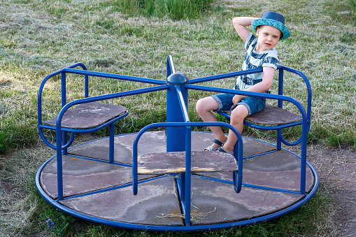 A children's merry-go-round is spinning in the playground and a cute boy is sitting and resting on it.
