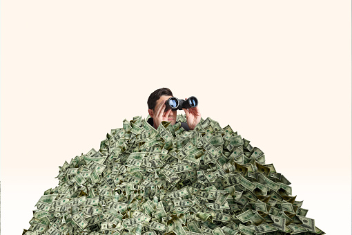 A man who is  buried in a pile of cash looks through a pair of binoculars.