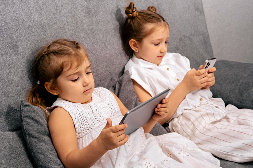Two girls sitting on sofa using modern gadgets to watch videos and play games.