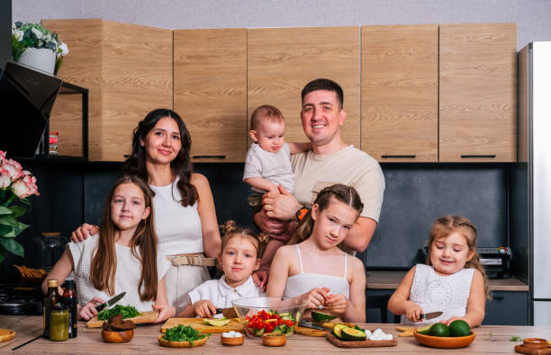 A large family - mom, dad, 4 daughters and a baby son together prepare a salad for lunch in a modern kitchen. Big family together concept. A large family - mom, dad, 4 daughters and a baby son together prepare a salad for lunch in a modern kitchen. Big family together concept. 3 6 months stock pictures, royalty-free photos & images