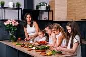 Young mother teaching her four daughters to cook green salad at the kitchen. Family together concept.