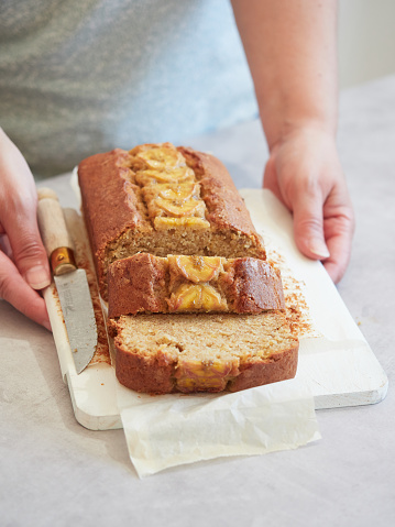 woman's hands holding freshly baked banana bread on white cutting board with baking paper and knife at the table.