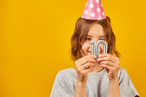 Cheerful, positive ten years birthday girl in party hat with candles, happy, on yellow studio background.