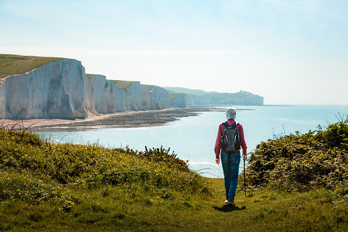 Color image depicting a senior man in his 70s hiking on a path leading down to the iconic Seven Sisters cliffs on the coastline of East Sussex, UK. The man is wearing casual clothing - blue denim jeans, a red checked shirt, navy blue gilet and a rucksack.