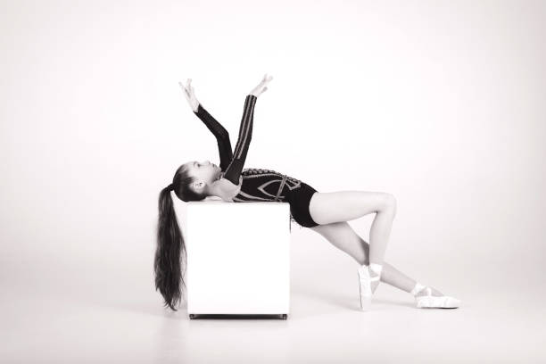 A beautiful girl gymnast in a black leotard does tricks on a white cube on a white background. White and black picture. Front view