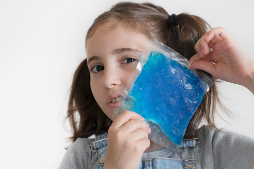 Little girl is applying blue ice gel pack to her chin in pain on white